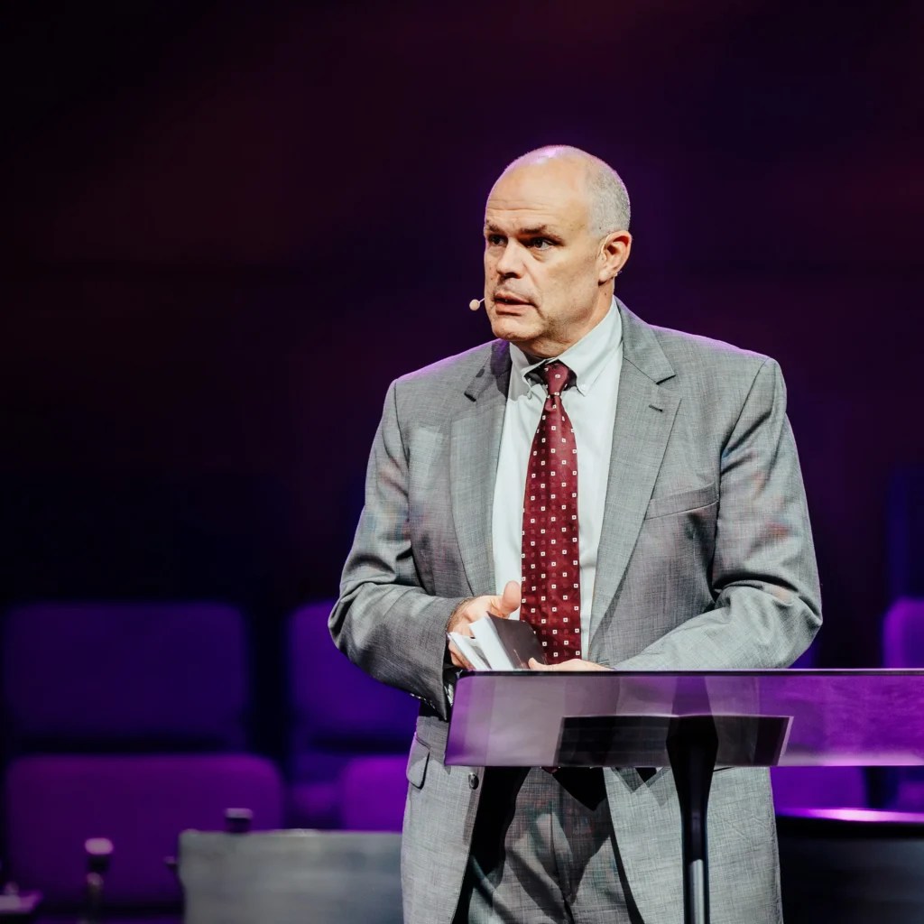Pastor David Rose speaking at the pulpit in a gray suit during his sermon at Trinity Baptist Church.