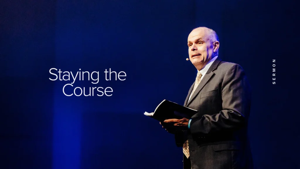 Pastor David Rose holding an open Bible on stage — Staying the Course sermon at Trinity Baptist Church.